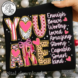 May include: Black graphic design with the words "YOU ARE" in a variety of patterns, including pink hearts, leopard print, and checkered designs. The words "Enough, Brave, Worthy, Loved, Amazing, Strong, Capable, Smart, Kind" are also displayed.