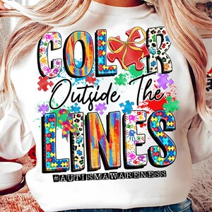 May include: White sweatshirt with a colorful graphic design that says "Color Outside The Lines" with puzzle pieces and the hashtag #autismawareness.