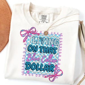 Pu&ograve; includere: T-shirt color crema con la scritta "LIVING ON THAT Blue Collar DOLLAR" in un design blu e rosa. Il disegno &egrave; incorniciato da un motivo a scacchi blu e bianco e fiocchi rosa. Una collana d'oro &egrave; drappeggiata sulla maglietta.