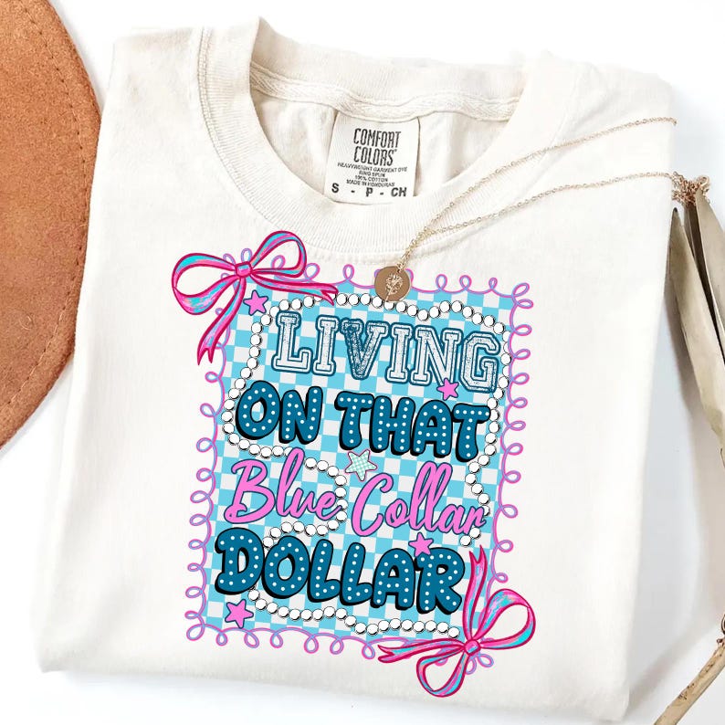 Pu&ograve; includere: T-shirt color crema con la scritta "LIVING ON THAT Blue Collar DOLLAR" in un design blu e rosa. Il disegno &egrave; incorniciato da un motivo a scacchi blu e bianco e fiocchi rosa. Una collana d'oro &egrave; drappeggiata sulla maglietta.