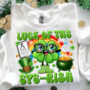 May include: White sweatshirt with a green four-leaf clover wearing glasses and holding an eye chart. The clover is smiling and has a rainbow behind it. The text "Luck of the Eye-rish" is written in green letters.