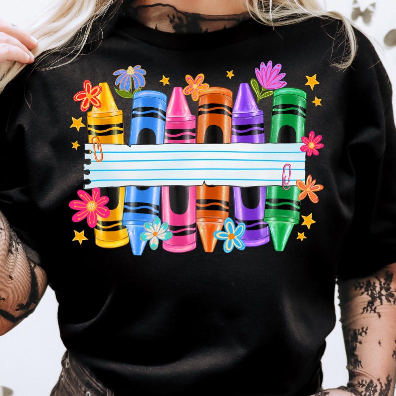 May include: Black t-shirt featuring a colorful crayon design. The design includes yellow, blue, pink, orange, purple, and green crayons, along with flowers and stars. A lined paper banner is in the center.