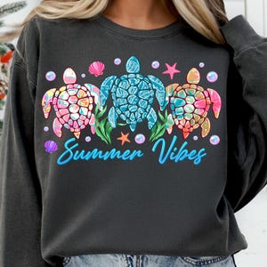 May include: A dark gray sweatshirt with a graphic of three colorful sea turtles and the text "Summer Vibes".