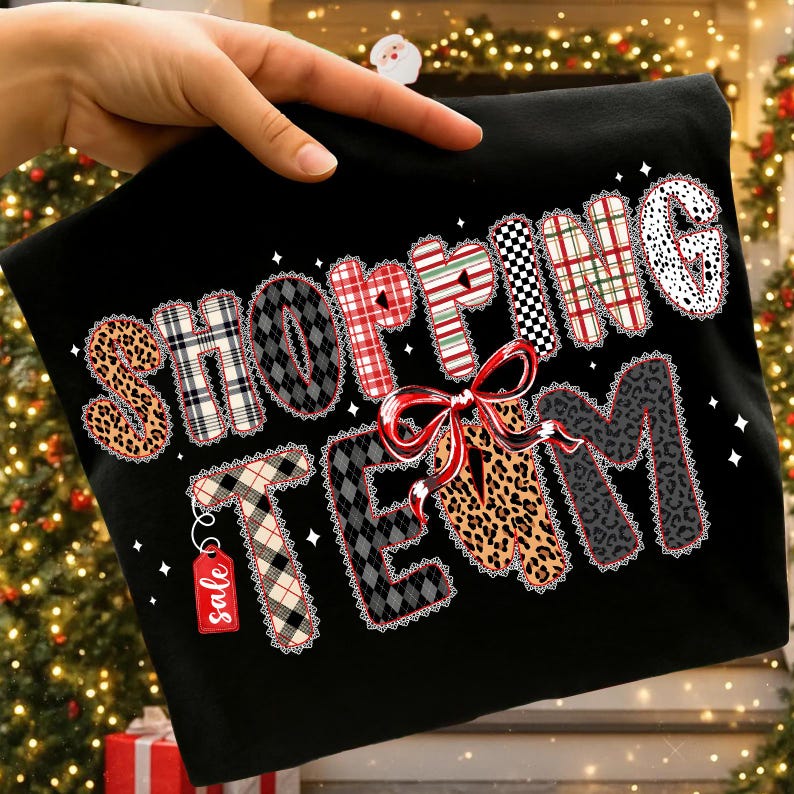 Peut inclure: T-shirt noir avec l'inscription "SHOPPING TEAM" dans diff&eacute;rents motifs, dont l&eacute;opard, tartan et vichy. Un n&oelig;ud rouge et une &eacute;tiquette "soldes" sont &eacute;galement pr&eacute;sents. Le t-shirt est tenu par une main.