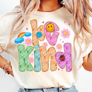 May include: Cream-colored sweatshirt with the words "be kind" in colorful, outlined letters. The design includes a rainbow, flowers, smiley faces, and a ladybug. The graphic is in a retro, groovy style.