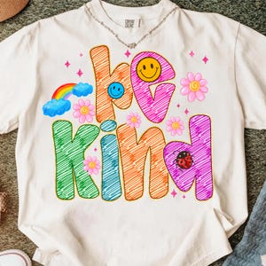 May include: Cream-colored t-shirt with the words "be kind" in colorful, whimsical lettering. The design includes a rainbow, flowers, a ladybug, and smiley faces. The shirt is made of a soft material.