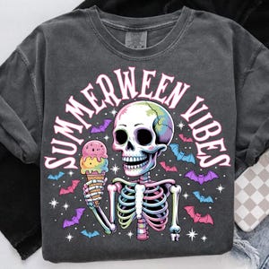 May include: Dark gray t-shirt with a skeleton graphic holding an ice cream cone. The design includes the text "SUMMERWEEN VIBES" in pink, along with colorful bats and stars. The skeleton's skull has rainbow coloring.