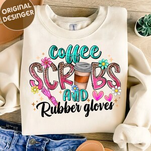 May include: A cream-colored sweatshirt with the text "Coffee Scrubs and Rubber Gloves" in pink, blue, and green lettering. The text is surrounded by flowers, hearts, and a coffee cup.