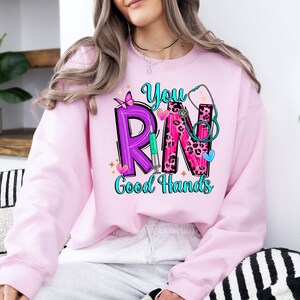 May include: Pink sweatshirt with a graphic design of the letters "RN" in purple and pink with a stethoscope, butterfly, and syringe. The text "You" is above the letters and "Good Hands" is below.