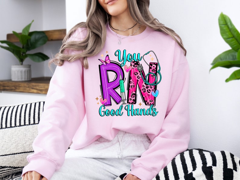 May include: Pink sweatshirt with a graphic design of the letters "RN" in purple and pink with a stethoscope, butterfly, and syringe. The text "You" is above the letters and "Good Hands" is below.