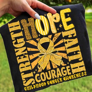 May include: Black t-shirt with gold text and a yellow ribbon design. The shirt features the words "Strength, Hope, Faith, Courage" and "Childhood Cancer Awareness". The word "Hope" has a leopard print design.