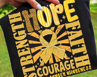 May include: Black t-shirt with gold text and a yellow ribbon design. The shirt features the words "Strength, Hope, Faith, Courage" and "Childhood Cancer Awareness". The word "Hope" has a leopard print design.