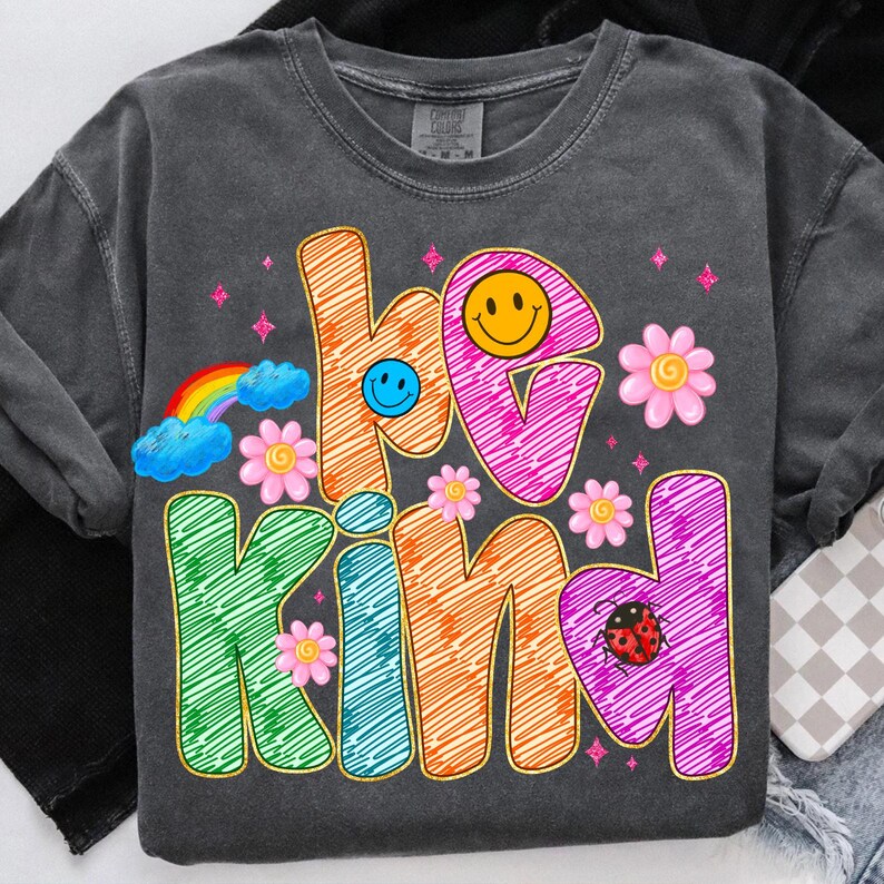 May include: Dark gray sweatshirt with the words "be kind" in colorful, scribbled letters. The design includes a rainbow, smiley faces, flowers, and a ladybug, creating a cheerful and positive vibe.