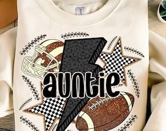 May include: Cream-colored sweatshirt featuring a football theme. The design includes a football helmet, a football, and two stars with a checkered pattern. The word "auntie" is in the center, with a black and gray lightning bolt.