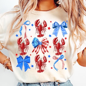以下が含まれることがあります： クリーム色のTシャツ。赤いロブスター、青いリボン、赤と青の星の模様。リボンは赤と白のストライプと青い星の愛国的なデザイン。独立記念日の祝賀に最適です。