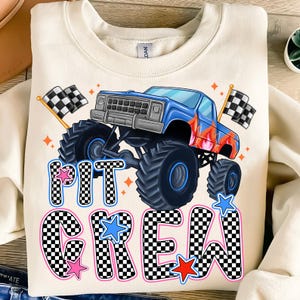 May include: A white sweatshirt with a blue monster truck graphic and the text "PIT CREW" in black and white checkered font with pink, red, and blue stars.