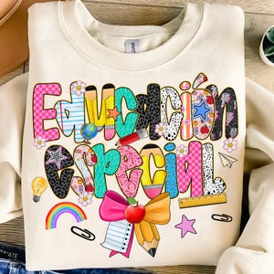 Pode incluir: Camisola creme com as palavras "Educación Especial" em letras coloridas e caprichosas. O design inclui gráficos temáticos escolares como lápis, maçãs e um arco-íris. Um tema divertido e educativo.