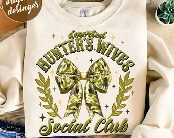 Op de afbeelding: Crèmekleurige sweatshirt met de tekst "deserted HUNTER'S WIVES Social Club" in groen en goud. Een camouflage strik is het centrale ontwerpelement, omringd door een lauwerkrans en sterren. De onderkant van de afbeelding luidt "300 DPI SUBLIMATION DESIGNS PNG FILE".