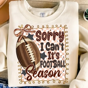 May include: Cream-colored sweatshirt with a football graphic and the text "Sorry I Can't It's Football Season." The graphic includes a brown football, stars, and a decorative border. A bow is at the top left of the graphic.