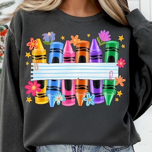 May include: Dark gray sweatshirt featuring a colorful crayon design. The design includes crayons in various colors, flowers, stars, and a blank lined paper. A paper clip is attached to the paper. The sweatshirt is a comfortable, casual style.