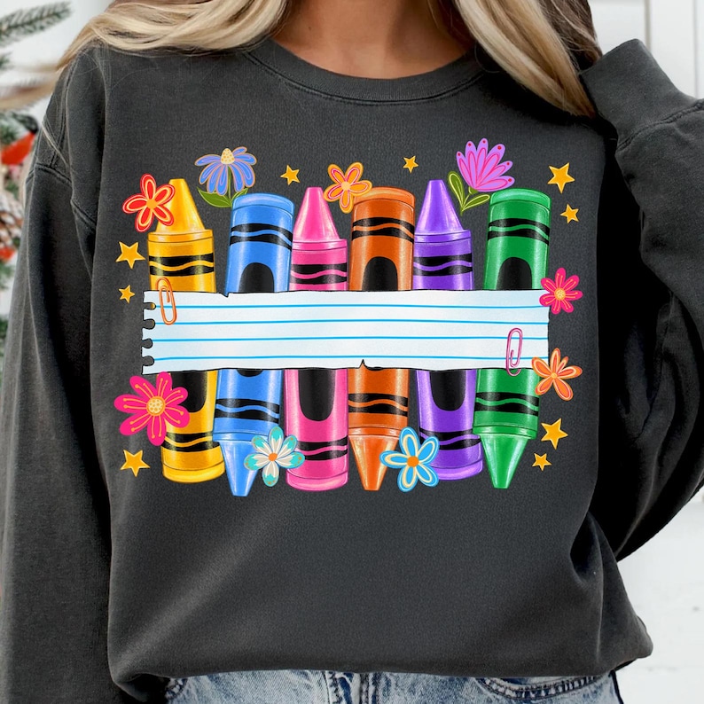 May include: Dark gray sweatshirt featuring a colorful crayon design. The design includes crayons in various colors, flowers, stars, and a blank lined paper. A paper clip is attached to the paper. The sweatshirt is a comfortable, casual style.