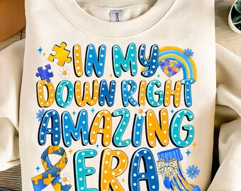 May include: A white sweatshirt with a blue, yellow, and white design that says "In My Down Right Amazing Era". The design includes a puzzle piece, a rainbow, a heart, and a ribbon.