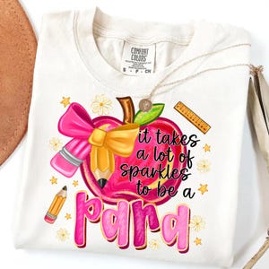 May include: Cream-colored t-shirt with a colorful graphic. The design features a pink apple with a bow and pencil, and the text "it takes a lot of sparkles to be a para". A gold necklace is draped on the shirt.