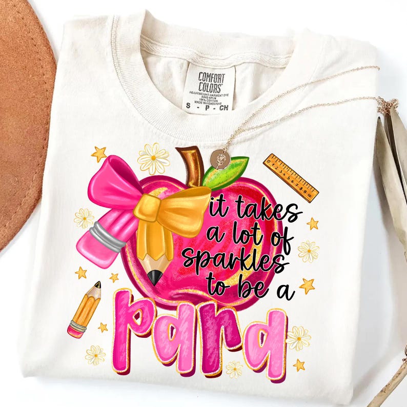 May include: Cream-colored t-shirt with a colorful graphic. The design features a pink apple with a bow and pencil, and the text "it takes a lot of sparkles to be a para". A gold necklace is draped on the shirt.