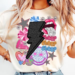 May include: Cream-colored sweatshirt with a graphic design featuring a black lightning bolt, pink lips, and the word "Cheer" in multiple colors. Other graphics include stars, hearts, and a disco ball.