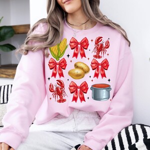 K&ouml;nnte beinhalten: Ein rosa Sweatshirt mit einem festlichen Design mit roten Schleifen, Maiskolben, Kartoffeln, einem Hummer und einem Topf mit roter Sauce.