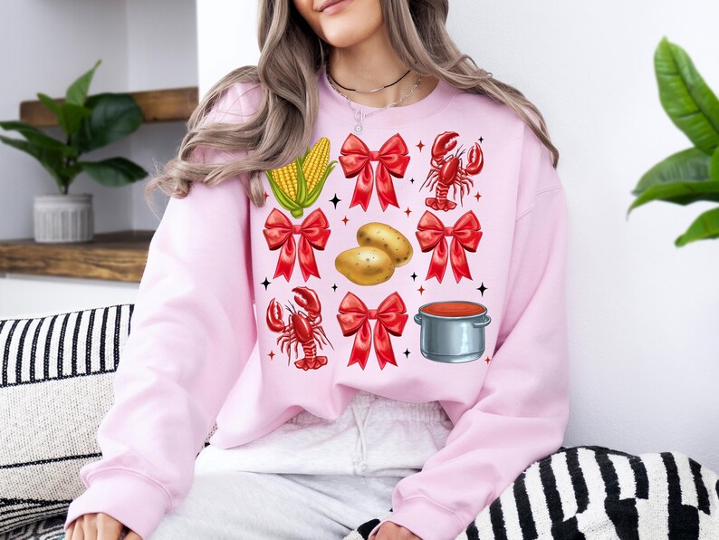 K&ouml;nnte beinhalten: Ein rosa Sweatshirt mit einem festlichen Design mit roten Schleifen, Maiskolben, Kartoffeln, einem Hummer und einem Topf mit roter Sauce.