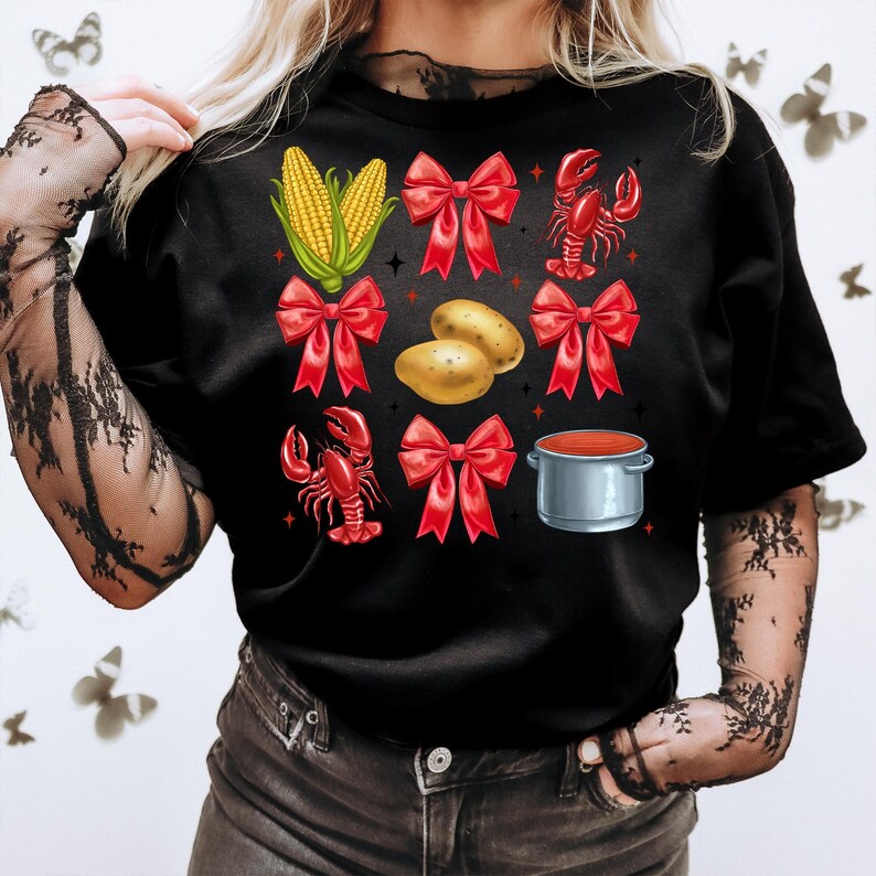 K&ouml;nnte beinhalten: Schwarzes T-Shirt mit einem rot-wei&szlig;-gr&uuml;nen Design mit einem Krebs, Mais auf der Kolbe, Kartoffeln und einem Topf mit roter Sauce. Das Design wird mehrmals auf dem Shirt wiederholt.