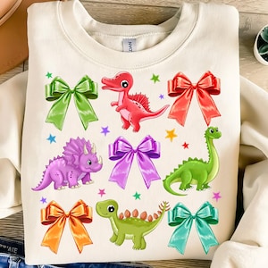 May include: A white sweatshirt with a colorful design featuring dinosaurs and bows. The dinosaurs are green, red, and purple, and the bows are green, red, purple, orange, and blue.