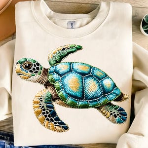 May include: A white sweatshirt with an embroidered sea turtle design. The turtle is green, blue, and brown with a detailed shell.