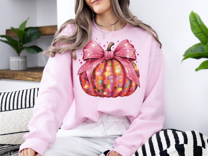 May include: A light pink sweatshirt featuring a colorful pumpkin design. The pumpkin has a mosaic pattern in shades of orange, pink, and green, topped with a large pink bow. The design includes sparkling star accents.