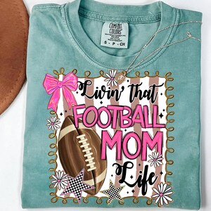 May include: A light teal Comfort Colors t-shirt with a football and the text "Livin' that Football Mom Life" in pink and black. The design includes a pink bow, stars, and flowers.
