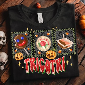 K&ouml;nnte beinhalten: Schwarzes T-Shirt mit einem Halloween-Motiv. Das Shirt zeigt drei quadratische Bilder mit einem Lutscher, einer Rose und einem Dessert. Das Wort "TRICOTRI" ist in Rot und Gr&uuml;n geschrieben. Maisbonbons und K&uuml;rbislaternen sind ebenfalls enthalten.