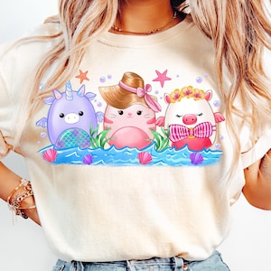 K&ouml;nnte beinhalten: Ein wei&szlig;es T-Shirt mit einem Aufdruck von drei Comic-Tieren im Meer. Die Tiere sind ein Einhorn, eine Katze mit Hut und ein Schwein mit einer Blumenkrone. Die Tiere l&auml;cheln alle und haben rosafarbene Wangen.