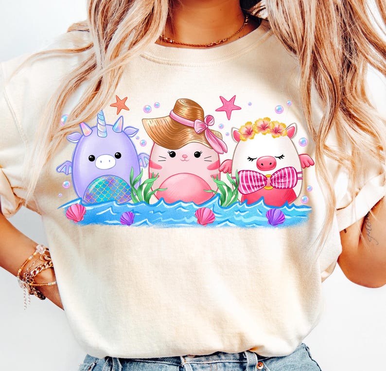 K&ouml;nnte beinhalten: Ein wei&szlig;es T-Shirt mit einem Aufdruck von drei Comic-Tieren im Meer. Die Tiere sind ein Einhorn, eine Katze mit Hut und ein Schwein mit einer Blumenkrone. Die Tiere l&auml;cheln alle und haben rosafarbene Wangen.