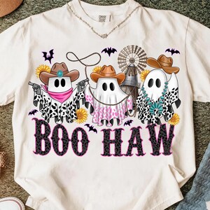 May include: Cream-colored t-shirt featuring three ghost characters in cowboy hats and bandanas, holding a lasso and guns. The phrase "BOO HAW" is printed in black and pink. Sunflowers and bats are also in the design.