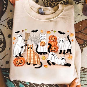 May include: Beige sweatshirt with a Halloween design. The illustration shows cartoon cats in ghost costumes, pumpkins, and plaid outfits. Bats, spiderwebs, and autumn leaves are also featured in the design.