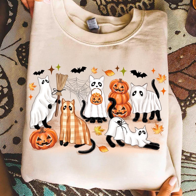 May include: Beige sweatshirt with a Halloween design. The illustration shows cartoon cats in ghost costumes, pumpkins, and plaid outfits. Bats, spiderwebs, and autumn leaves are also featured in the design.