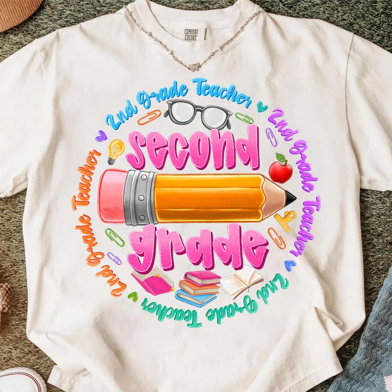 May include: Cream-colored t-shirt with a colorful graphic design for a 2nd Grade Teacher. The design includes a large pencil, the words "Second Grade" in pink, and other school-related icons like glasses, an apple, and books.