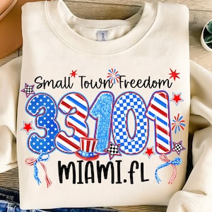 Puede incluir: Sudadera de color crema con un diseño patriótico. El gráfico incluye el texto "Small Town Freedom" y "33101 MIAMI.FL" en rojo, blanco y azul. Presenta estrellas, fuegos artificiales y un sombrero de copa.