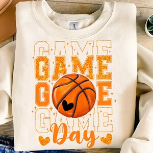 Peut inclure: Un sweat-shirt blanc avec le texte "Game Day" en lettres orange paillet&eacute;es. Un ballon de basket-ball de couleur orange et marron est au centre du texte. Le ballon de basket-ball a un c&oelig;ur noir au centre.
