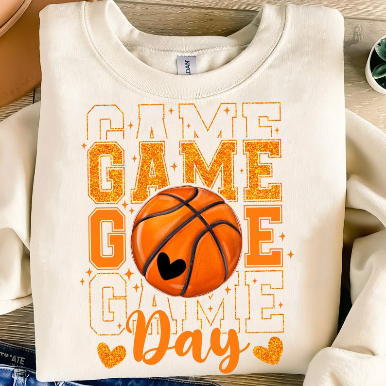 Peut inclure: Un sweat-shirt blanc avec le texte "Game Day" en lettres orange paillet&eacute;es. Un ballon de basket-ball de couleur orange et marron est au centre du texte. Le ballon de basket-ball a un c&oelig;ur noir au centre.