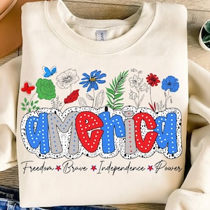 May include: A cream-colored sweatshirt with a graphic design of the word "america" in blue, red, and gray with white polka dots. The word is surrounded by colorful flowers and leaves. The text "Freedom Brave Independence Power" is printed below the graphic.