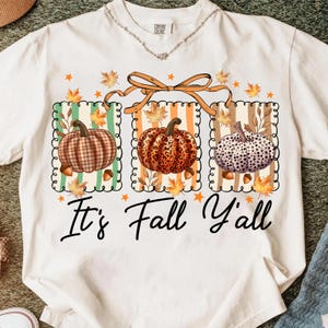 May include: White t-shirt featuring a fall-themed graphic with three pumpkins in different patterns. The pumpkins are set against striped backgrounds with fall leaves and the text "It's Fall Y'all" in a script font.