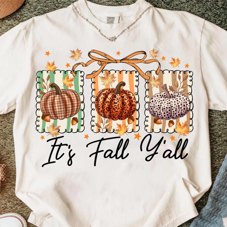 May include: White t-shirt featuring a fall-themed graphic with three pumpkins in different patterns. The pumpkins are set against striped backgrounds with fall leaves and the text "It's Fall Y'all" in a script font.
