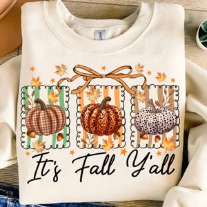 May include: Cream-colored sweatshirt featuring three decorative panels with pumpkins, leaves, and acorns. Each panel has a different pumpkin design and a striped background. The text "It's Fall Y'all" is written in a script font.